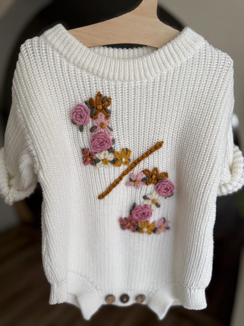 May include: A white knit baby bodysuit with short sleeves, featuring embroidered floral accents in pink, yellow, and brown. The bodysuit has a round neckline and three wooden buttons at the bottom. The garment is hanging on a wooden hanger.