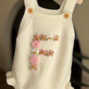 May include: Cream-colored knitted baby romper with a floral embroidered "F" on the front. The romper has wooden button closures and a ruffled hem. The garment is displayed on a wooden hanger.