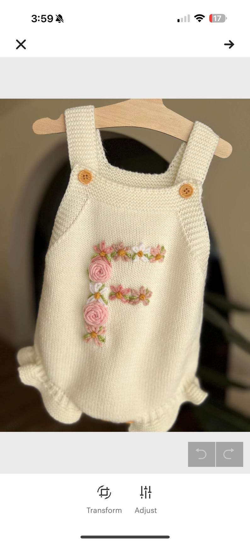 May include: Cream-colored knitted baby romper with a floral embroidered "F" on the front. The romper has wooden button closures and a ruffled hem. The garment is displayed on a wooden hanger.