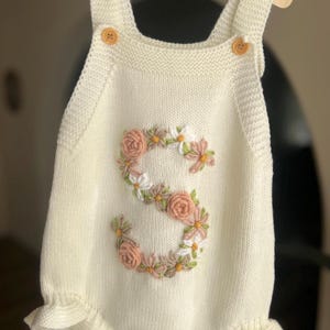 May include: Cream-colored knitted romper with a floral embroidered "S" design. The romper features wooden buttons on the straps and a ruffled hem. The floral embroidery includes pink and white flowers.