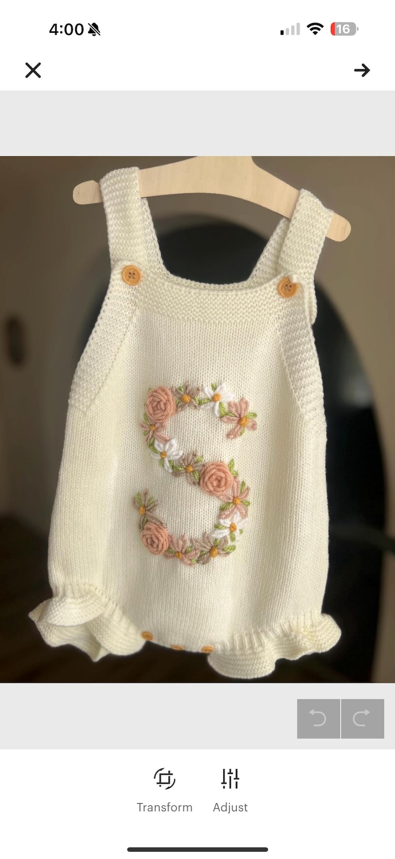 May include: Cream-colored knitted romper with a floral embroidered "S" design. The romper features wooden buttons on the straps and a ruffled hem. The floral embroidery includes pink and white flowers.