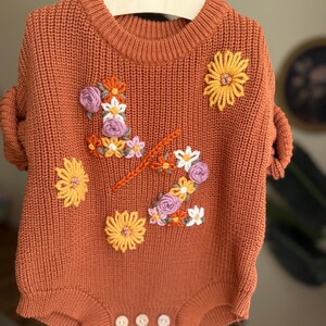 May include: Rust-colored baby romper with floral embroidery. The knit romper features a crew neck, short sleeves, and three buttons at the bottom. The floral embroidery includes yellow, white, and purple flowers, and an orange stem.