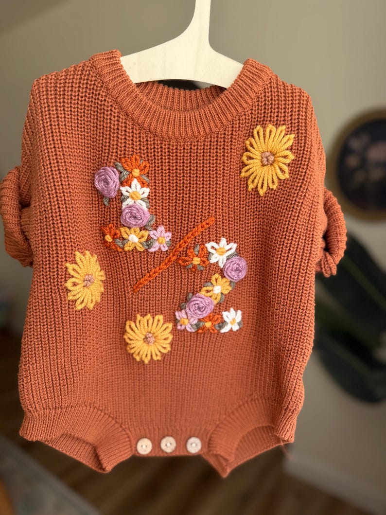May include: Rust-colored baby romper with floral embroidery. The knit romper features a crew neck, short sleeves, and three buttons at the bottom. The floral embroidery includes yellow, white, and purple flowers, and an orange stem.