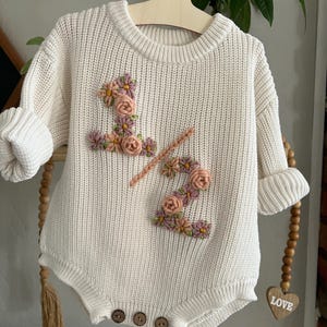 May include: Cream-colored baby romper with embroidered floral "1/2" design. The romper features a round neckline, long sleeves with rolled cuffs, and wooden buttons. The floral embroidery includes pink, peach, and yellow flowers. The romper is hanging on a wooden hanger.