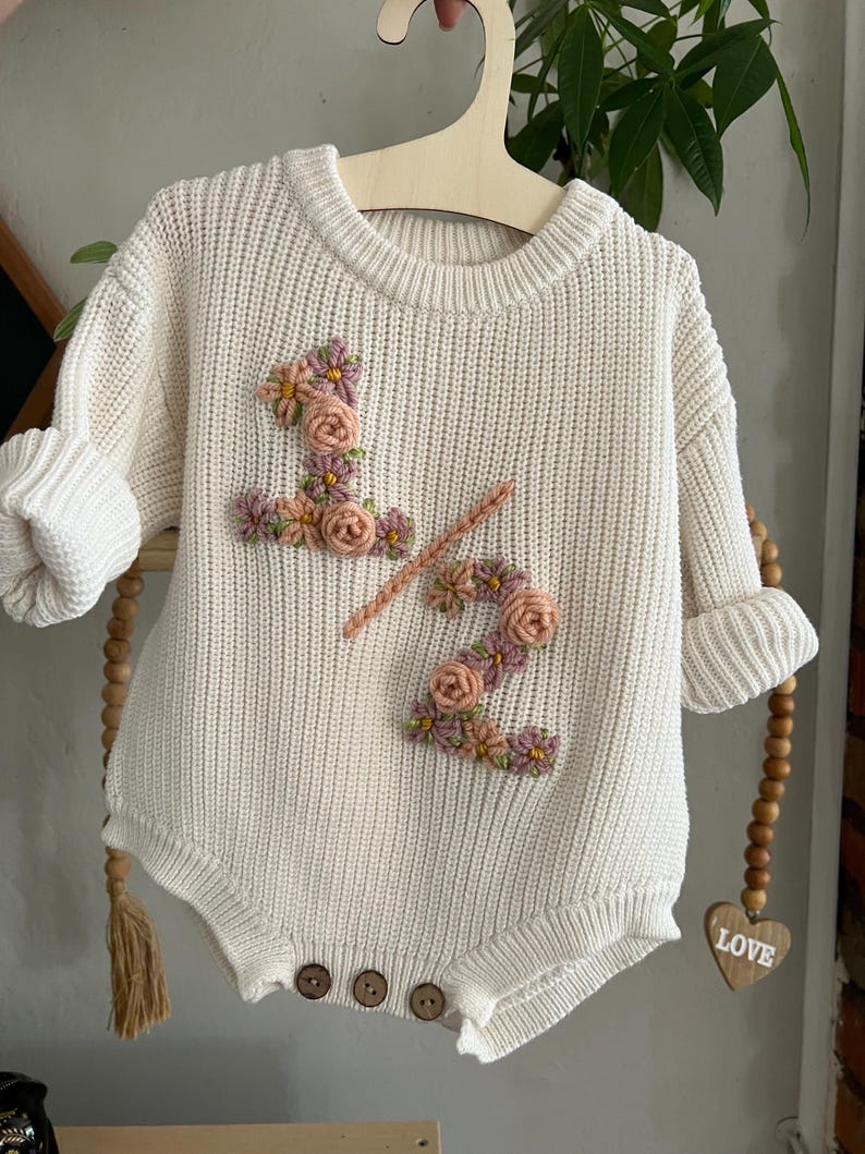 May include: Cream-colored baby romper with embroidered floral "1/2" design. The romper features a round neckline, long sleeves with rolled cuffs, and wooden buttons. The floral embroidery includes pink, peach, and yellow flowers. The romper is hanging on a wooden hanger.