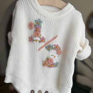 May include: Cream-colored knitted baby romper with floral embroidery. The design features the number "1/2" adorned with pink, blue, and white flowers. The romper has a round neck, long sleeves, and three brown buttons at the bottom.