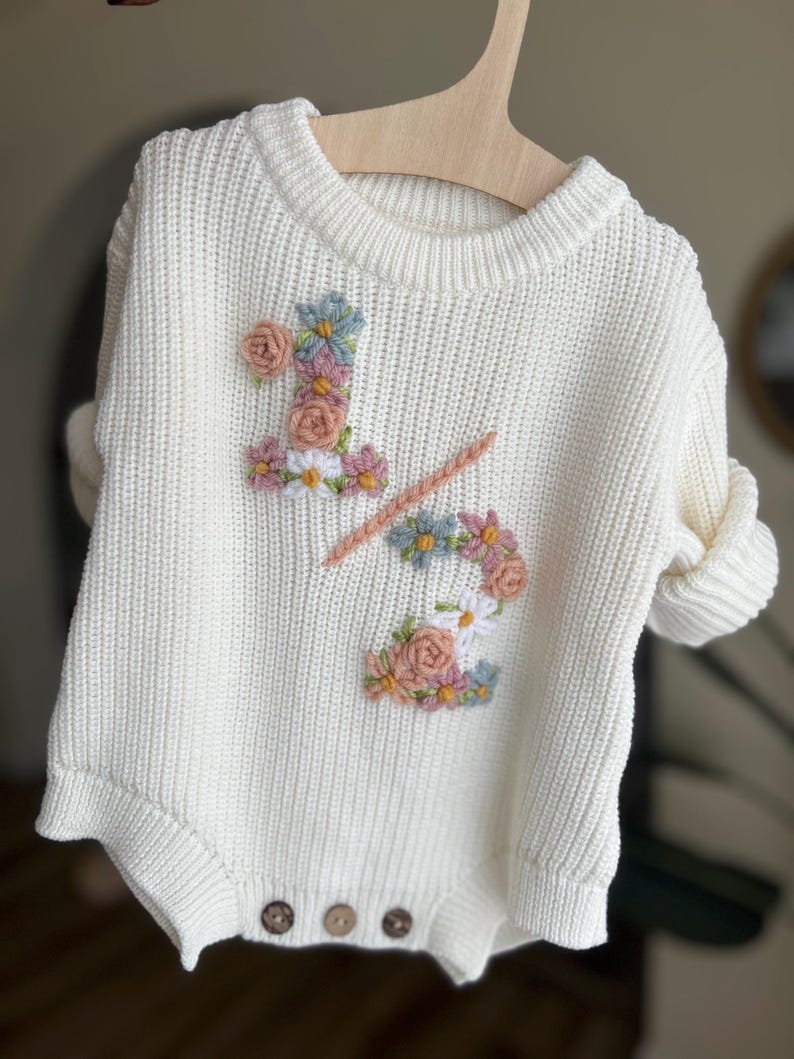 May include: Cream-colored knitted baby romper with floral embroidery. The design features the number "1/2" adorned with pink, blue, and white flowers. The romper has a round neck, long sleeves, and three brown buttons at the bottom.