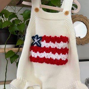 May include: White knitted baby romper with a red, white, and blue crocheted American flag design on the front.