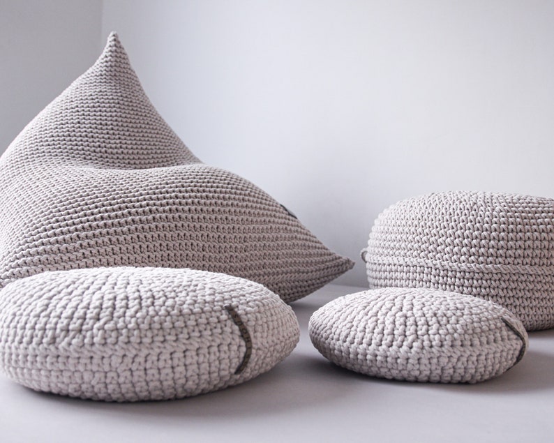 May include: A set of four beige knitted poufs, including a large, triangular pouf and three smaller round poufs. The poufs are made of a soft, textured fabric and are perfect for adding a touch of comfort and style to any room.