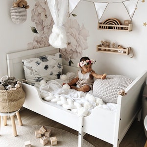 May include: A white toddler bed with a white crocheted blanket and a grey knitted ottoman. A baby girl wearing a floral headband sits on the bed with a wooden rainbow toy on a shelf behind her. The room is decorated with a white floral wall decal and a white lace canopy.