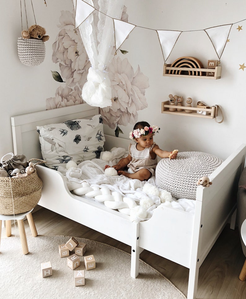 May include: A white toddler bed with a white crocheted blanket and a grey knitted ottoman. A baby girl wearing a floral headband sits on the bed with a wooden rainbow toy on a shelf behind her. The room is decorated with a white floral wall decal and a white lace canopy.