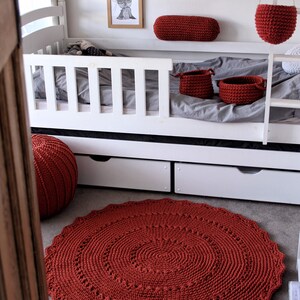 May include: A red crocheted rug with a circular design, placed on a gray floor in front of a white bunk bed. The bed has a red crocheted pillow and two red crocheted baskets.