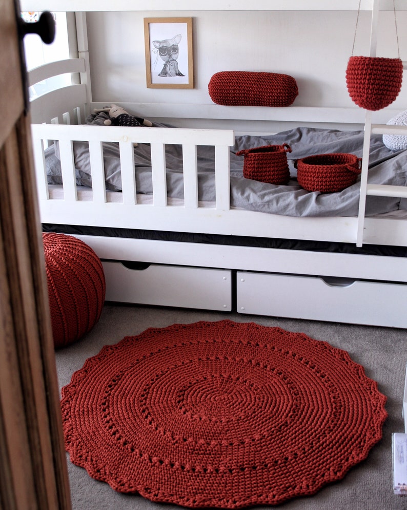 May include: A red crocheted rug with a circular design, placed on a gray floor in front of a white bunk bed. The bed has a red crocheted pillow and two red crocheted baskets.
