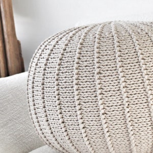 May include: A large, round, beige knitted cushion with a textured surface. The cushion is sitting on a white sofa.