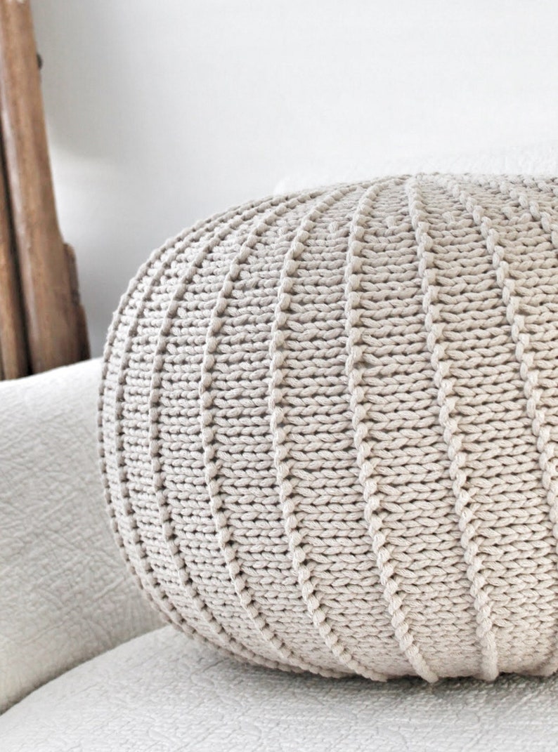 May include: A large, round, beige knitted cushion with a textured surface. The cushion is sitting on a white sofa.