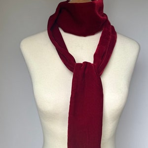 May include: A luxurious deep red velvet scarf displayed on a mannequin. The scarf is knotted at the front, with long, flowing ends. The fabric has a soft texture and the colour is a rich, saturated hue. A classic accessory.