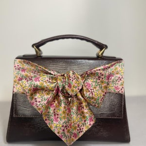 May include: A brown leather handbag with a floral patterned bow. The bag has a top handle and a flap closure.