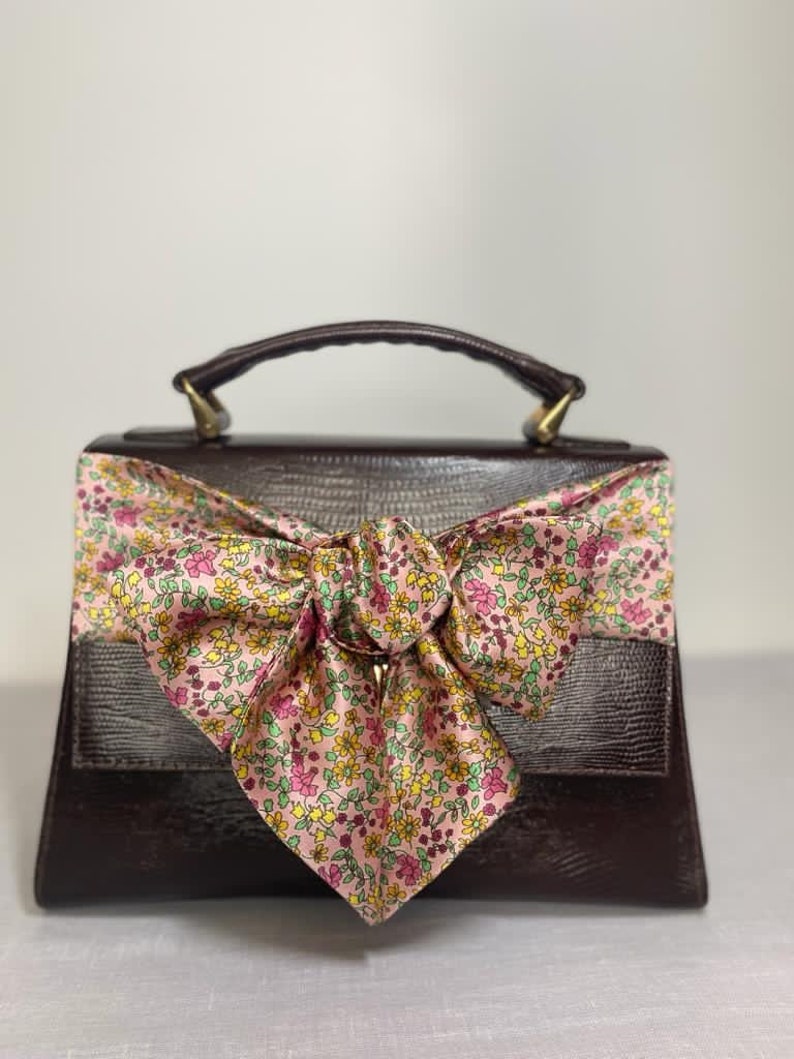 May include: A brown leather handbag with a floral patterned bow. The bag has a top handle and a flap closure.