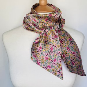 May include: A pink satin scarf with a floral print in shades of pink, yellow, and green. The scarf is tied around a wooden mannequin's neck.