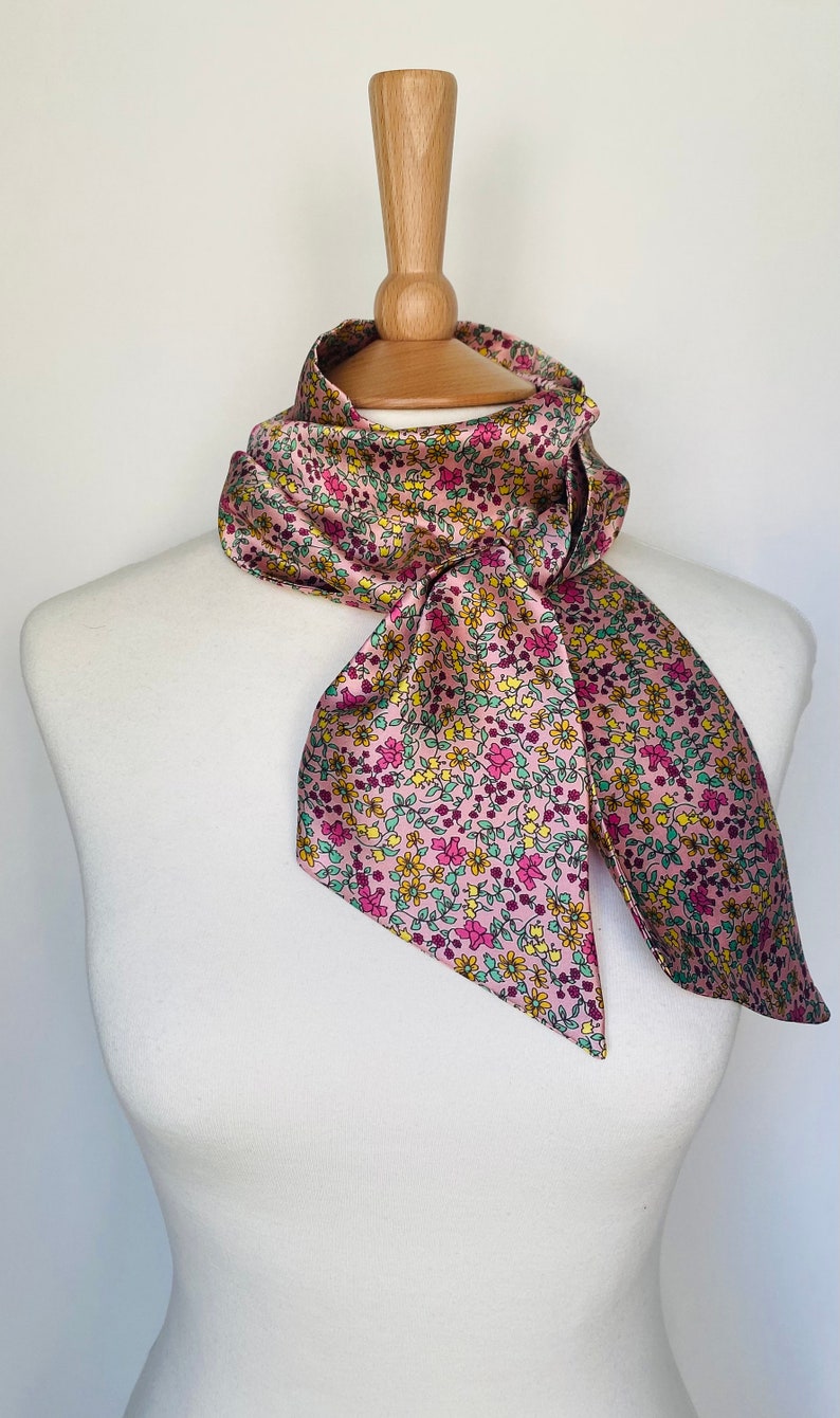 May include: A pink satin scarf with a floral print in shades of pink, yellow, and green. The scarf is tied around a wooden mannequin's neck.