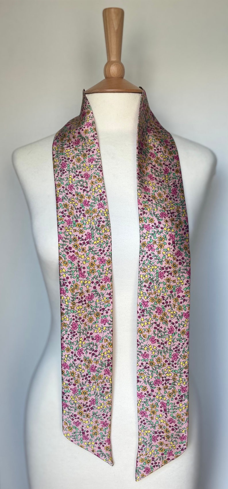 May include: A long, pink scarf with a floral pattern. The scarf has small pink, yellow, and green flowers on a light pink background.