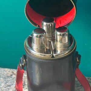May include: A black cylindrical carrying case with a red interior lining and a zip closure, containing three silver-toned metal flasks with rounded caps. The case features a matching black and red shoulder strap attached to the sides.