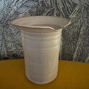 May include: A beige ceramic pitcher with a speckled texture and a wide, flared rim. The vessel features a cylindrical body with subtle horizontal ridges along the exterior surface and a small, integrated pouring spout on one side of the rim.