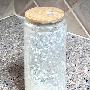 May include: A clear glass jar with a white glitter and star design. The jar has a wooden lid with a hole in the centre.