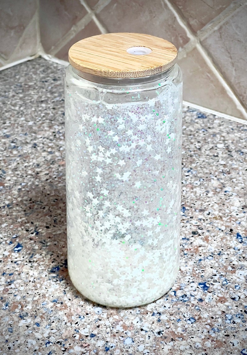 May include: A clear glass jar with a white glitter and star design. The jar has a wooden lid with a hole in the centre.