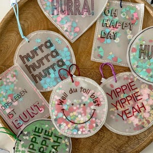 Op de afbeelding: Een verzameling heldere, cirkelvormige en hartvormige tags met kleurrijke confetti en tekst. De tags bevatten zinnen als "HURRA!", "OH HAPPY DAY" en "YIPPIE YIPPIE YEAH". Elke tag heeft een gekleurd touwtje om op te hangen.