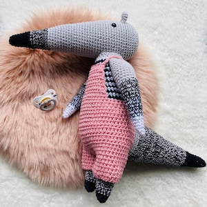 May include: A handmade crochet anteater doll with a gray head and tail, black and white striped arms, and pink overalls. The doll has black feet and a pacifier is next to it. The doll is sitting on a pink fluffy surface.