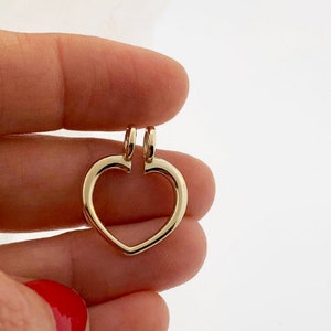 May include: A gold-colored heart-shaped pendant with two small loops at the top for a chain. The pendant is held between two fingers against a white background. The heart has a smooth, polished finish.