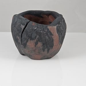 May include: A handmade, small, round ceramic bowl with a rough, textured exterior. The bowl is primarily black with brown and tan accents. The interior is a warm brown color. The bowl has a cracked design on the side.