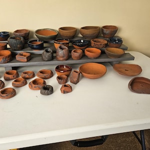 May include: A collection of handmade ceramic bowls and containers in various sizes and shapes. The pottery features earthy tones of brown, orange, and blue, with textured surfaces and unique designs. Some pieces are stacked on a tiered display.