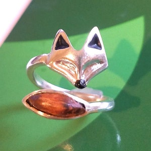 Puede incluir: Un anillo de plata con un diseño de zorro. La cabeza del zorro está mirando hacia adelante con ojos negros y una cola enroscada.