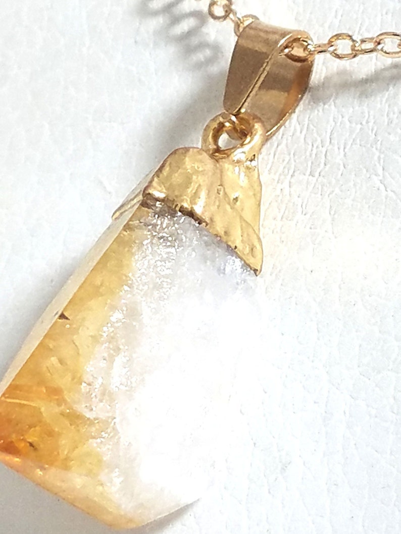 K&ouml;nnte beinhalten: Eine goldene Halskette mit einem Anh&auml;nger aus einem rohen, nat&uuml;rlichen Citrin-Kristall mit einem wei&szlig;en, wolkigen Inneren. Der Kristall ist in einer goldenen Fassung gefasst und h&auml;ngt an einer zarten goldenen Kette.