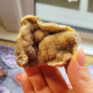 Può includere: Un campione minerale grezzo di colore marrone chiaro con una struttura cristallina. La roccia ha una consistenza irregolare ed è tenuta in una mano. Il minerale sembra essere una geode o una formazione rocciosa simile.