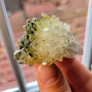 Puede incluir: Un grupo de cristales de cuarzo transparente con una base amarillo-verdosa. El cuarzo se sostiene en una mano, con una ventana y una escena exterior en el fondo. Los cristales tienen un aspecto natural y brillante.