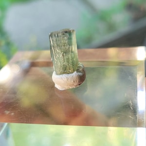 May include: A green and gray crystal on a clear glass display stand. The crystal is a rectangular prism with a pointed top.