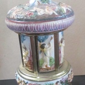 May include: A porcelain carousel music box featuring a conical lid topped with a small cherub figurine. The central rotating section displays relief panels of cherubs playing instruments, framed by brass pillars, set on a brass base with cherubs.