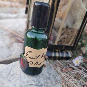 May include: A small, green glass bottle with a black cap and a handwritten label that reads "Enuit herb Oil." The bottle has a decorative, colorful embellishment. The background includes a decorative lantern and natural elements.