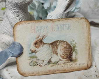 May include: A vintage-style Easter card with a brown and white rabbit illustration. The card is decorated with a blue and white ribbon and has the text "Happy Easter" printed on it. The card is numbered "No. 26".
