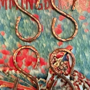May include: Four copper-colored, hammered metal S-hooks. The hooks are arranged in a square pattern against a background of red, green, and blue colors. The hooks have a textured surface and are designed for hanging items.