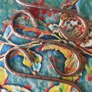May include: Three copper-colored S-hooks with a hammered texture are displayed against a colorful, patterned background. The hooks are curved and have a rustic, handmade appearance, suitable for hanging items.