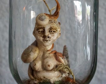 May include: A sculpture of a humanoid figure with snail shell details, inside a glass jar. The figure has a light beige complexion, with brown and beige accents. The clear jar contains natural elements at the base, creating a miniature scene.