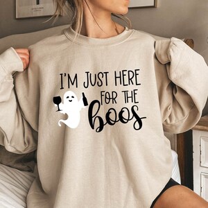 Pode incluir: Camisola bege com o texto preto "I'M JUST HERE FOR THE boos". Um fantasma de desenho animado segurando uma taça de vinho e uma garrafa é impresso na frente. A camisola é um estilo casual e confortável.