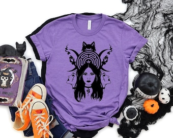 Puede incluir: Una camiseta de color morado jaspeado con un estampado gráfico negro de una figura de tres caras con un símbolo de laberinto, una silueta de lobo y lunas crecientes. El diseño incluye una cara central flanqueada por dos perfiles.