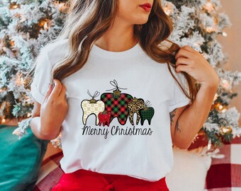 May include: White t-shirt featuring a festive design of teeth shaped like Christmas ornaments. The ornaments have various patterns, including gold filigree, red plaid, and leopard print. The words "Merry Christmas" are written below the ornaments.