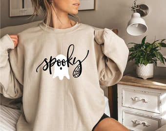 Könnte beinhalten: Beiges Sweatshirt mit dem Wort "spooky" in schwarzer Schrift, begleitet von einem weißen Geist und einem Spinnennetz-Design. Das langärmlige Sweatshirt ist perfekt für Halloween.