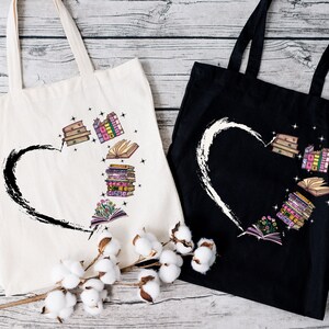 May include: Two tote bags, one cream and one black, with a heart design. The heart is formed by a black brushstroke on the cream bag and a white brushstroke on the black bag. Inside the heart are illustrations of books in various colors.
